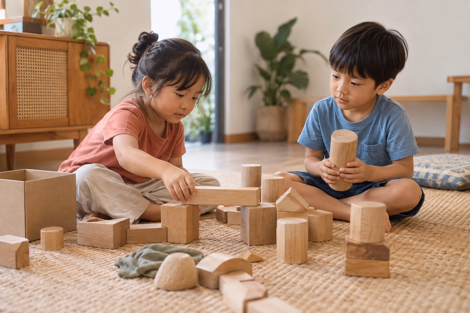Two children build a low bridge with large wooden blocks on a woven mat, with a cardboard box beside them.
